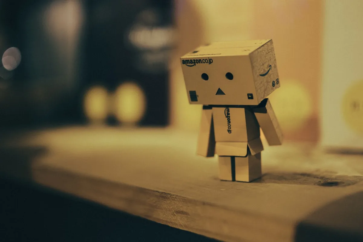 A small cardboard robot figurine sitting on a wooden shelf in a warmly lit room.
